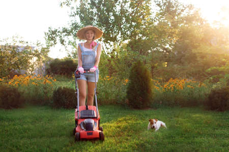 Women gardener. A sexy girl in a hat mows the grass near the house with a lawnmower. Landscape design and lawn careの写真素材