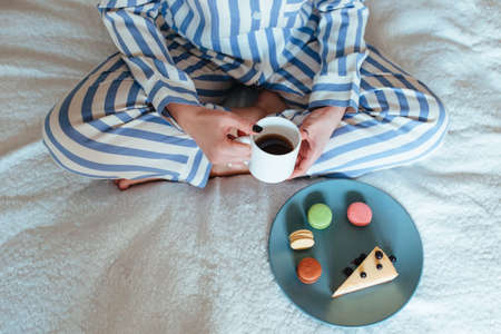 a woman in the morning drinks coffee and eats sweets cheesecake sweet macaroni. breakfast in bed conceptの写真素材