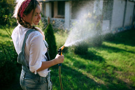 a young woman in a white shirt and denim overalls watering the lawn. gardening concept and landscape designの写真素材