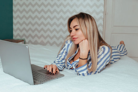 a beautiful girl in pajamas is lying on the bed behind a laptop. the concept of remote work at home and shoppingの写真素材