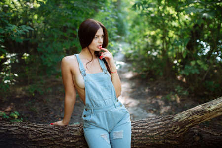 Young woman in jeans overalls standing under tree in woodlandの写真素材