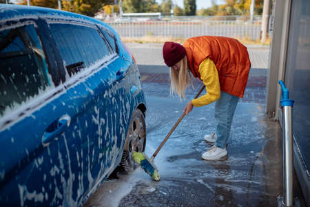 Beautiful young girl washes the car at the self-service car wash. erasing dirt to the car with a brushの写真素材