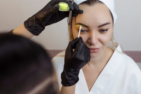 The hand of a make-up artist in black gloves applies a yellow eyebrow paste to the eyebrow....
