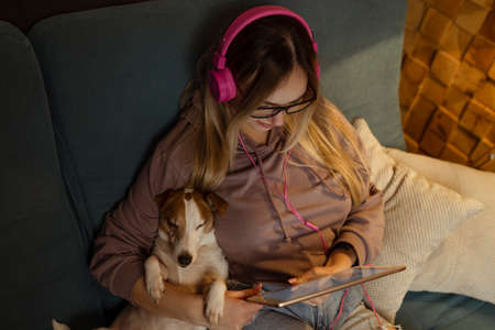 A girl on the couch lies with a dog in her laptop and listens to music through headphones. Freelancing and communication via the Internetの写真素材