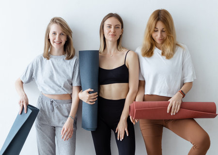 A group of young sports girls with yoga mats standing against a white wall. Girlfriends in the gym relaxing after fitness or yoga, indoorsの写真素材