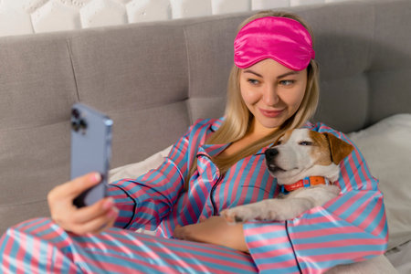 Portrait of beautiful joyful woman in pajamas hugging dog using mobile phone for selfie smiling while lying on bed after sleep or napの写真素材