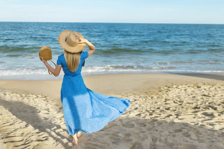 Woman in dress with straw hat walking by sea on sunny day, back viewの写真素材