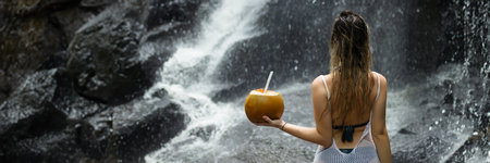 Beautiful woman stands facing a waterfall in Bali and holds a coconut in her handの写真素材