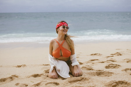 Young girl relaxes on the beach with white sand by the ocean. Beautiful model in a red swimsuit sunbathes. Blonde with curly hair in a bikini.の写真素材
