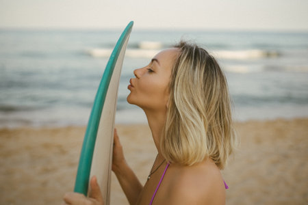 Blonde woman in purple bikini playfully kisses surfboard on sandy beach, summer sunset atmosphere, joyful and carefree vacation mood.の写真素材