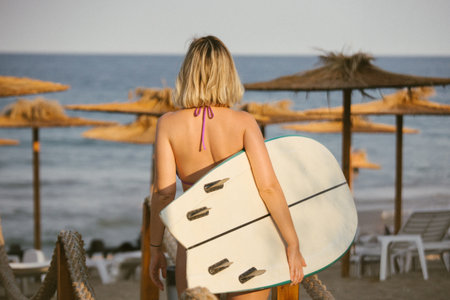 Young woman in bikini walking toward the sea with a surfboard, beach umbrellas in background, summer lifestyle and vacation mood, rear view, sunny evening light.の写真素材