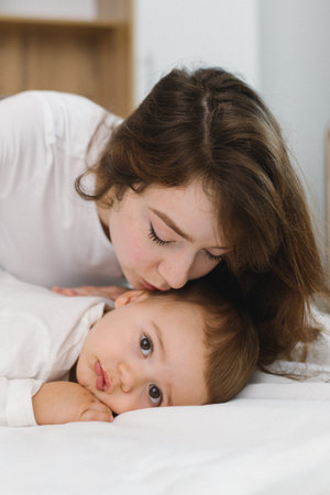 Cozy moment of a young woman and baby relaxing together at home, lying on a soft bed and smiling in...