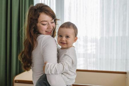 Cozy family moment with a young woman holding her smiling baby at home, standing near soft window light. A warm and cozy parent-child scene filled with loveの写真素材
