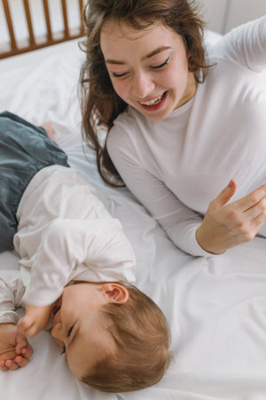 Happy mother and baby lying together on a soft white bed, enjoying a warm and cozy family moment filled with love, comfort, connection, and natural light.の写真素材