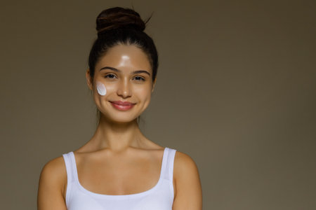 Young woman smiling with a skincare cream applied to her cheek, demonstrating a daily beauty routine.の写真素材