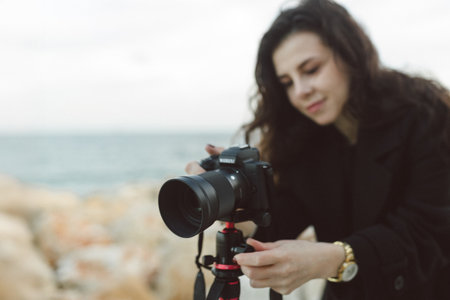 Female photographer adjusting professional camera on tripod outdoors by the sea, focusing the lens while preparing for a creative photo shoot on a rocky coastline.の写真素材