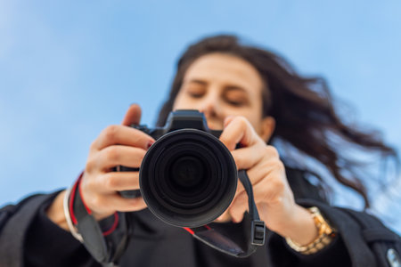 Low-angle view of a female photographer holding a professional camera against the blue sky, capturing outdoor moments and focusing on creative photography.の写真素材