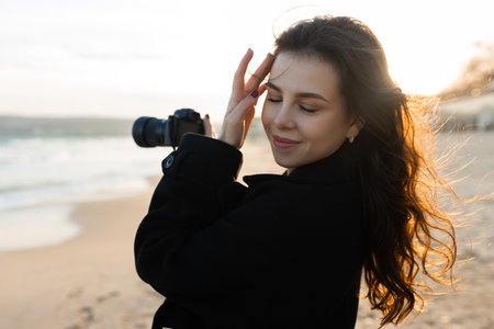 Smiling female photographer holding a camera on a sunny beach, enjoying golden hour light and capturing outdoor moments on the coastline.の写真素材