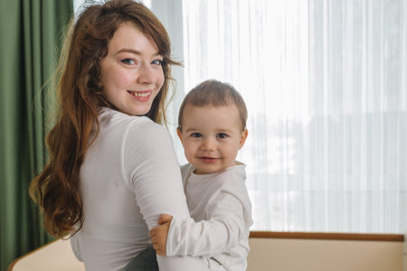 Cozy family moment with a young woman holding her smiling baby at home, standing near soft window...