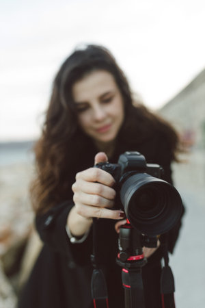 Female photographer adjusting professional camera on tripod outdoors by the sea, focusing the lens...