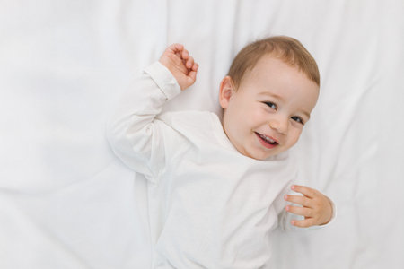 Cozy portrait of a happy baby lying on a soft white bed at home, smiling in natural light. A warm...