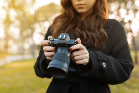 Close-up of a young woman holding a digital camera while photographing outdoors in warm sunlight....