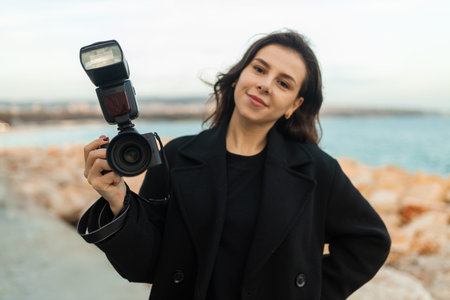 Young female photographer taking pictures outdoors by the sea, smiling while holding a professional...