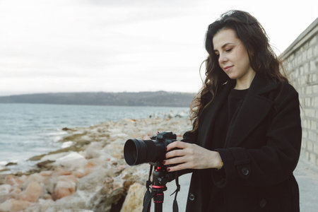 Female photographer adjusting professional camera on tripod outdoors by the sea, focusing the lens...