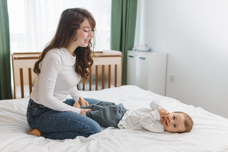 Young mother playing with her baby on a soft white bed, enjoying a warm and cozy family moment full...