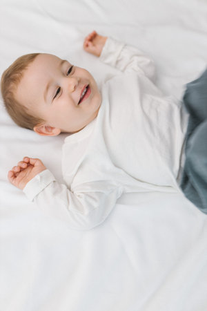 Cozy portrait of a happy baby lying on a soft white bed at home, smiling in natural light. A warm and cozy childhood moment filled with joy, innocence, comfort, and gentle family emotion.の写真素材