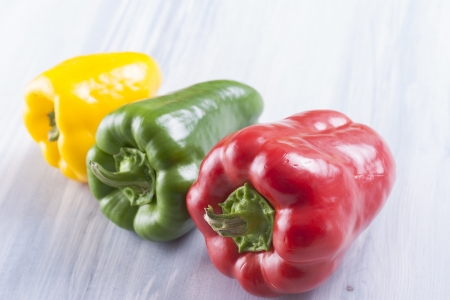 Close up photo of edible vegetables - a yellow, green and red bell pepper on a solid light blue wooden tableの写真素材