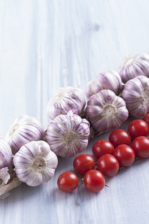 Close up photo of edible vegetables - a garlic bulbs and cherry tomatoes on a solid light blue wooden tableの写真素材