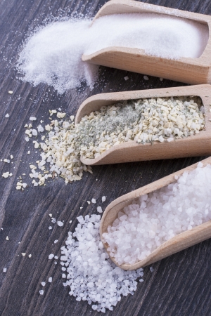Close up photo of a food ingredients in a wooden scoop - small pieces of a table salt, yellow colored garlic salt and large pieces of a sea salt placed on a dark wooden background.の写真素材
