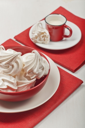 Side view of white meringues in a red bowl on a red textile napkin  In the distance are a red cup with milk and broken meringue on the white plate の写真素材