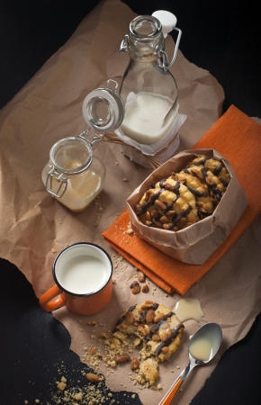 Top  view on the packaged peanut cookies, broken biscuit, cups and jars of milk on paper and a dark wooden background の写真素材