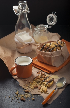 Side view on the packaged peanut cookies, broken biscuits, cups and jars of milk on paper and a dark wooden background の写真素材