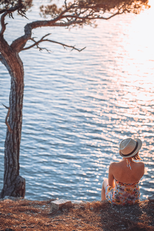 girl sitting by the sea at sunset, in a light summer dress and hat on the mountainside. She looks at the sea. Nearby stands a tree and the sun sets over the horizonの写真素材