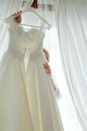 The bride is trying on a wedding dress, looking in front of her. She hangs on the coat rack with a dress of herself. In a bright key, behind the windowの写真素材