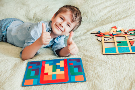 happy boy sits on the bed and lays puzzles. Child looks at the camera and shows thumbs up