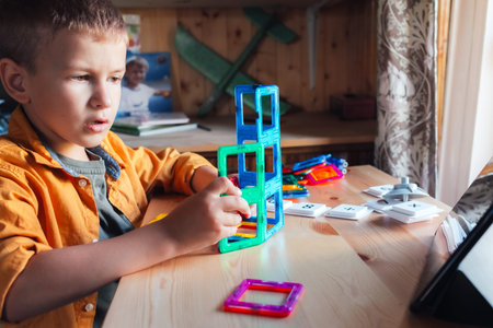 concentrated boy sits at desk and builds tower from magnetic constructor looking at the tabletの写真素材