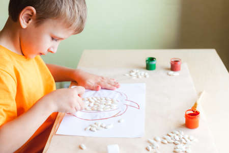 Happy boy making pumpkin picture from seeds on white paper, Halloween DIY concept. Step by step instruction. Step 2 Glue pumkin seeds onto the drawingの写真素材