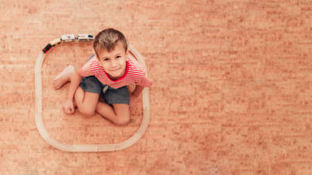 Cute little boy sitting on floor, around him is wooden railway with a train. Child looking at camera. Top view.の写真素材