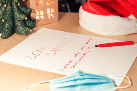 Desk with letter to Santa and decorated Christmas tree on background. Text Dear Santa please stop coronavirus on white paper. Medical mask lies nearbyの写真素材