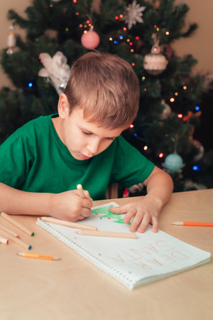 Smiling 7 years old boy wearing green t-short write letter to Santa sitting by desk, Christmas tree on backgroundの写真素材