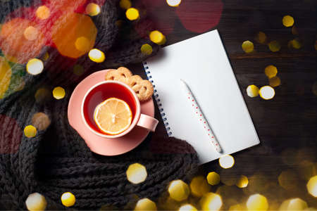 Clear copybook, cup of pomegranate tea, pen and knitted scarf on black wooden background with Christmas lightsの写真素材