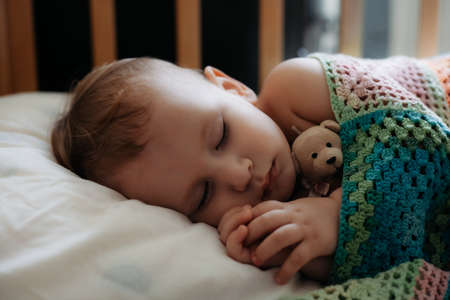 Cute baby sleeps peacefully in crib with handle under his cheek. One year old boy laying covered with DIY knitted blanketの写真素材