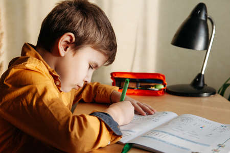 Cute 7 years old child doing his homework sitting by desk. Boy writing in notebook. Side viewの写真素材