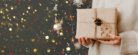 girl hands holding small gift in her hands. Close up. decorated Christmas tree on background. Bright...