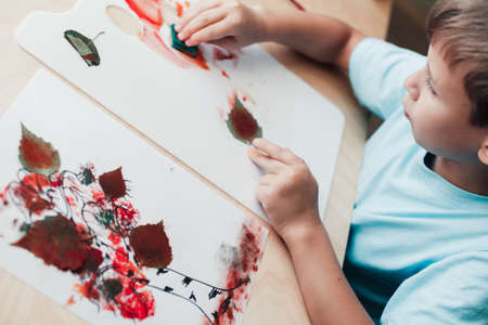 Cute child sitting at desk and making picture from dry birch leaves. Autumn activities for childrenの写真素材