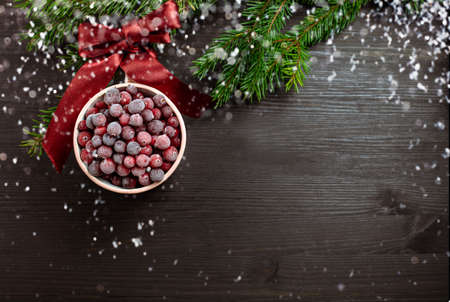 A cup with frozen cranberry berries on a dark wooden background looks like Christmas bauble. Branches of Christmas tree and snow. Flat lay with copy space on black wooden backgroundの写真素材